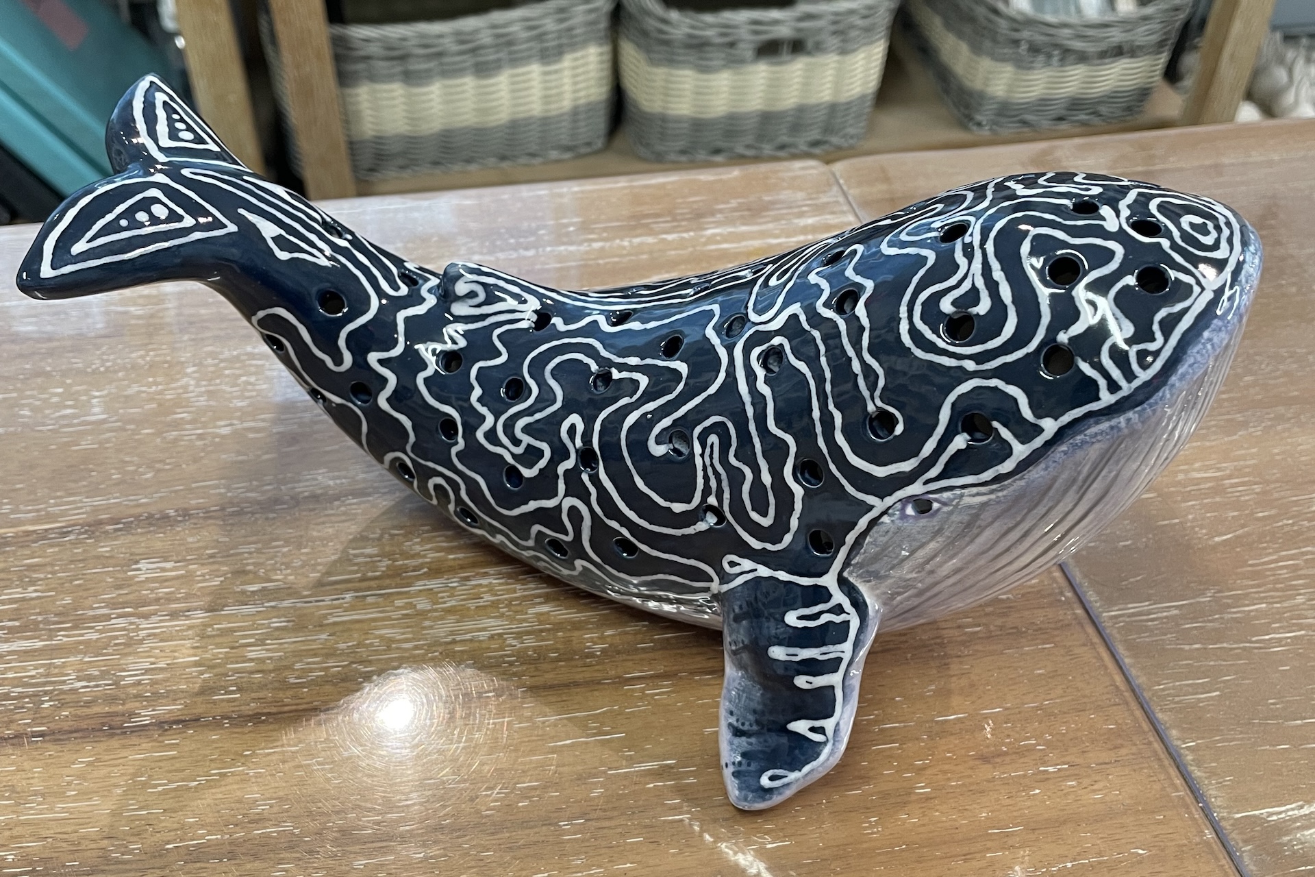 A ceramic whale figurine with intricate white patterns and small holes on its dark blue body, resting on a wooden surface with wicker baskets in the background.