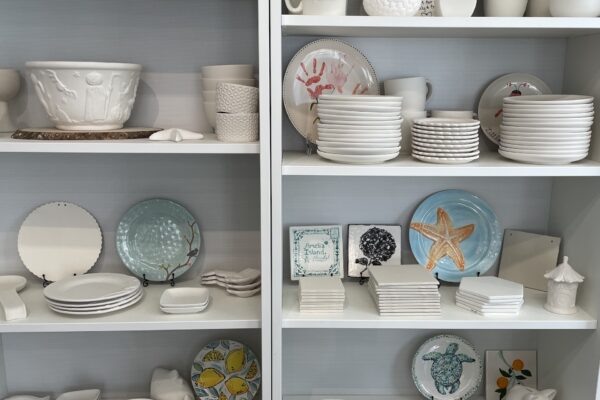 Shelves displaying assorted ceramic plates, bowls, mugs, and decorative items, some painted with colorful designs such as a starfish, tree, and sheep. The arrangement shows both plain and hand-painted pottery.