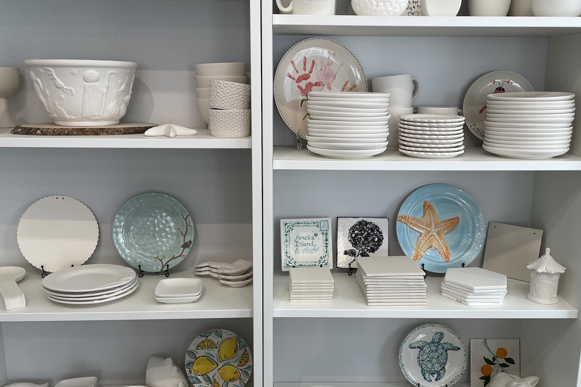 Shelves displaying assorted ceramic plates, bowls, mugs, and decorative items, some painted with colorful designs such as a starfish, tree, and sheep. The arrangement shows both plain and hand-painted pottery.