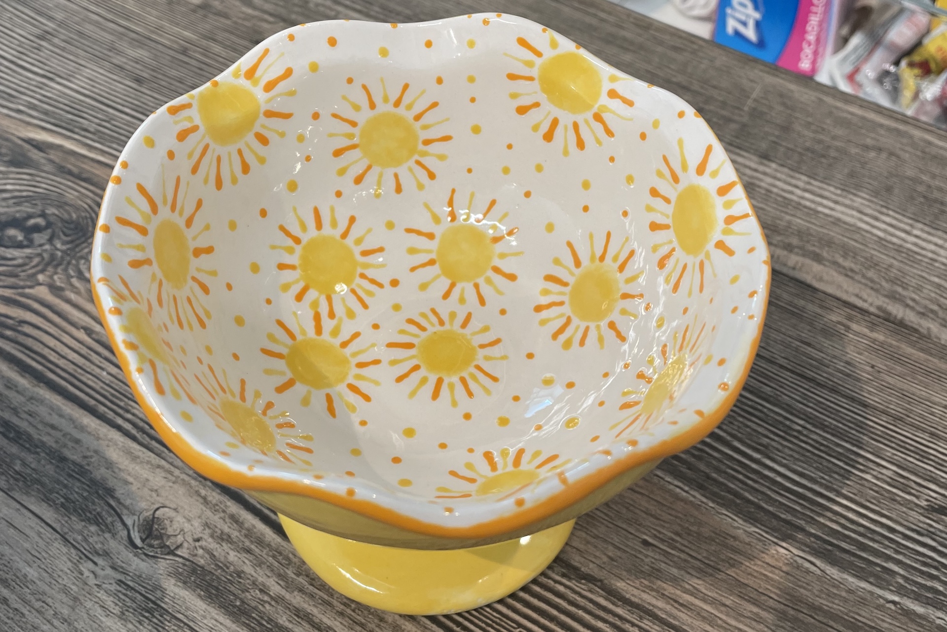 A ceramic bowl with a wavy rim, decorated with yellow sun patterns and dots on a white background, sits on a wooden surface. The bowl has a yellow base.