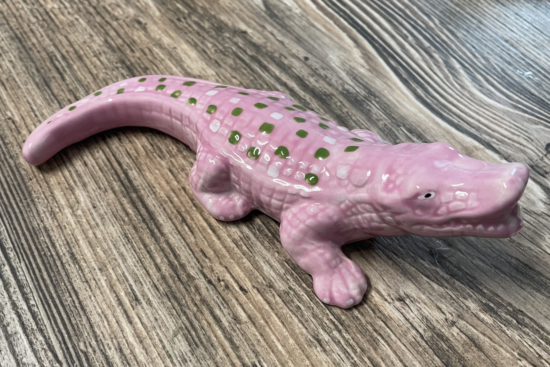 A ceramic figurine of a crocodile painted pink with green spots, displayed on a wood-grain surface. The crocodile has a slightly open mouth and visible texture on its body.