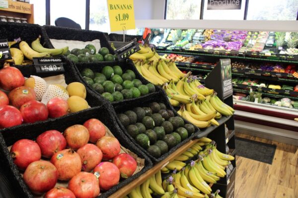 Produce section of a grocery store with pomegranates, avocados, mangos, and bananas in the foreground; various other fruits and vegetables are displayed on shelves in the background.