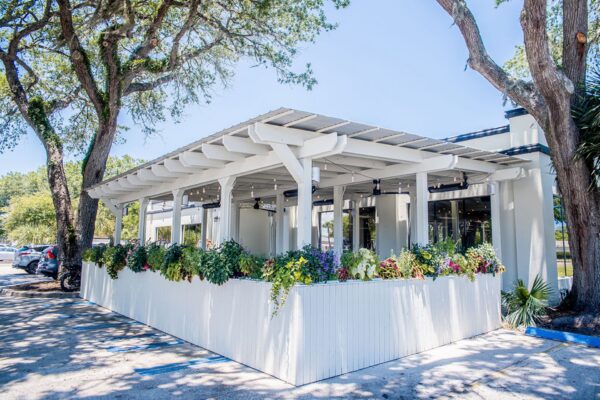 A white outdoor patio with a pergola, surrounded by lush green plants and colorful flowers, sits under tall trees on a sunny day.