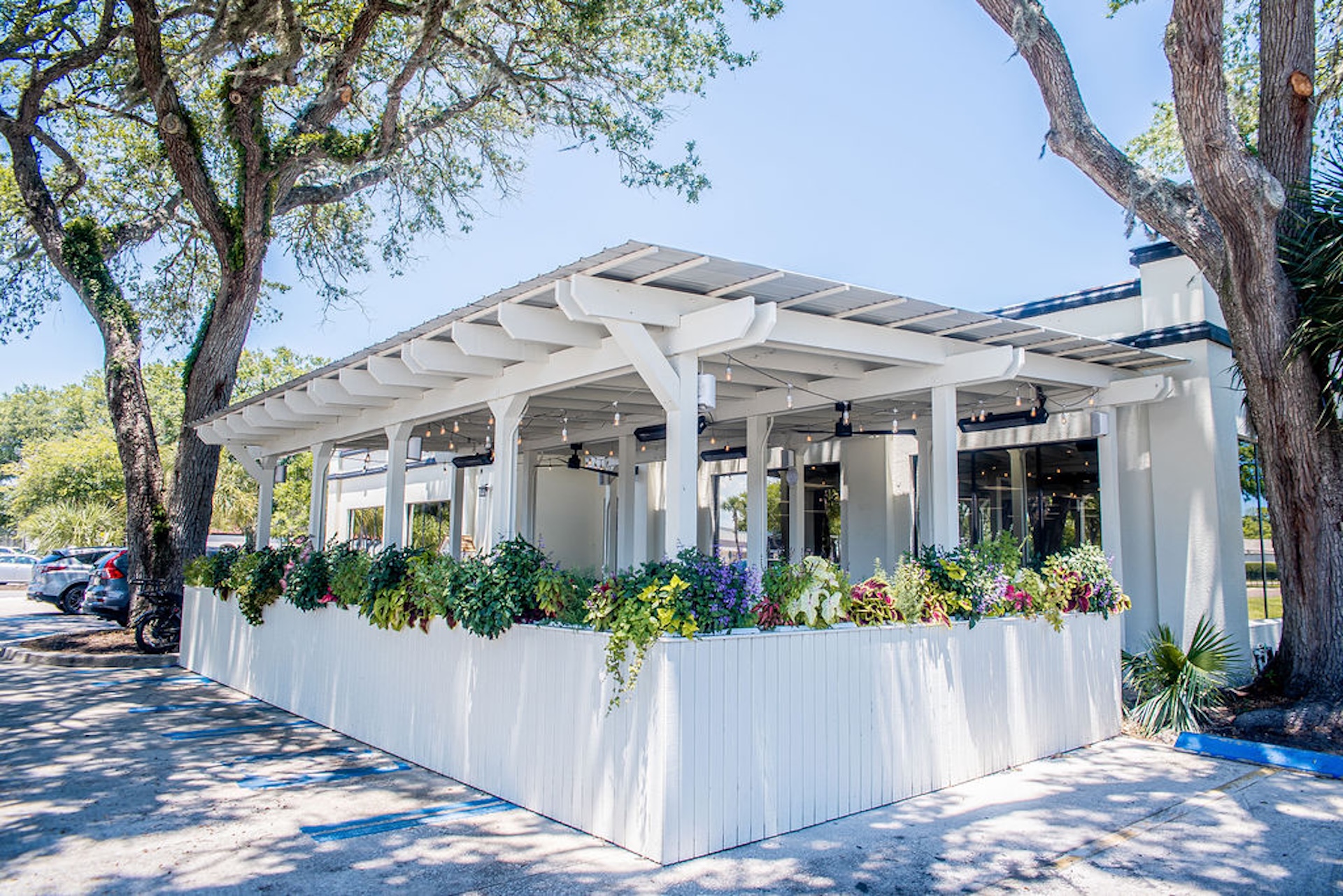 A white outdoor patio with a pergola, surrounded by lush green plants and colorful flowers, sits under tall trees on a sunny day.