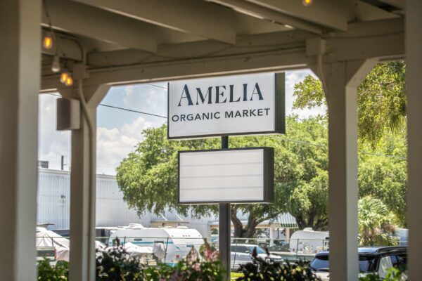 A white sign reads AMELIA ORGANIC MARKET outside, with trees, parked cars, and buildings in the background on a sunny day.