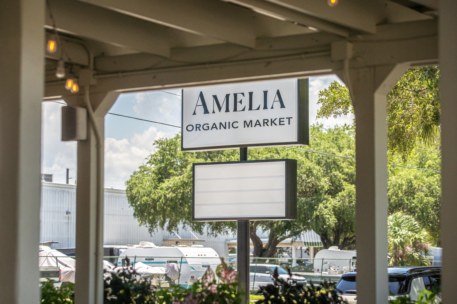 A white sign reads AMELIA ORGANIC MARKET outside, with trees, parked cars, and buildings in the background on a sunny day.
