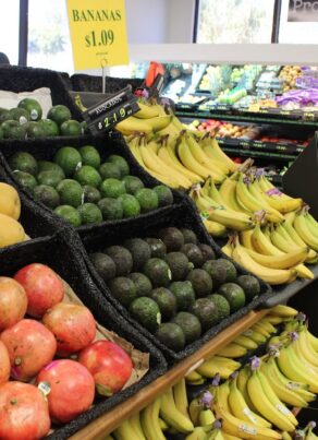 Grocery store produce section with bins of pomegranates, avocados, mangoes, and several bunches of bananas. Shelves in the background hold more fruits and vegetables.