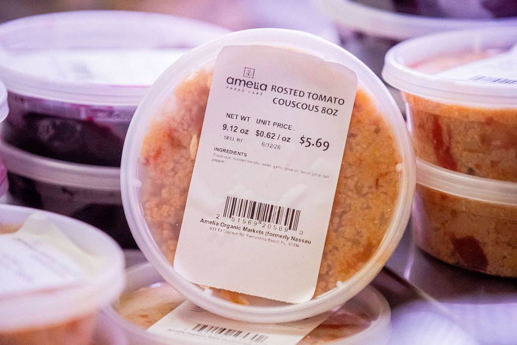 A plastic container labeled Rosted Tomato Couscous 8oz with a price of $5.69 sits on a shelf among other similar containers. The label also lists ingredients and nutritional information.