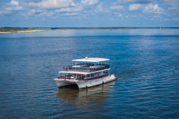 A white, double-deck tour boat sails on a calm blue body of water under a partly cloudy sky, with a distant shoreline and greenery visible in the background.