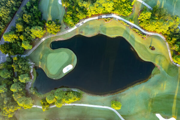 Aerial view of a golf course with a green island surrounded by a dark pond, winding paths, and lush green trees and fairways bathed in sunlight.