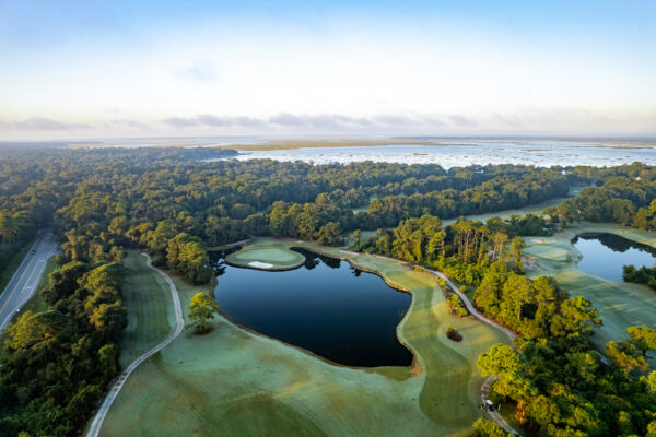 Aerial view of a golf course with green fairways, sand bunkers, ponds, and trees, bordered by a forest with a road on the left and water in the distance under a clear sky.