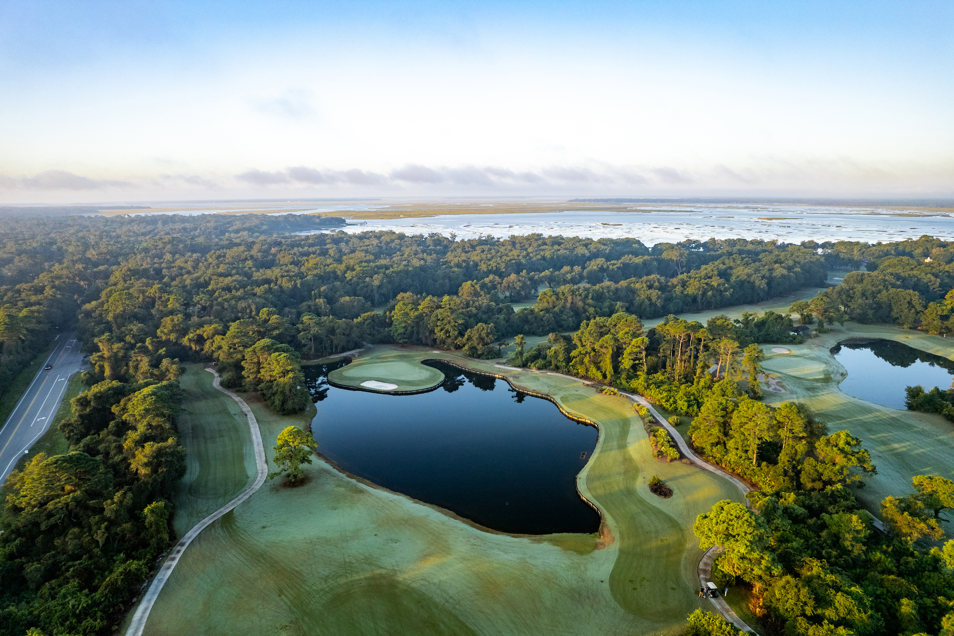 Aerial view of a golf course with green fairways, sand bunkers, ponds, and trees, bordered by a forest with a road on the left and water in the distance under a clear sky.