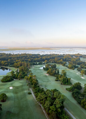 Aerial view of a lush, green golf course surrounded by trees and small ponds, with a road on the left and a distant body of water under a clear blue sky at sunrise.