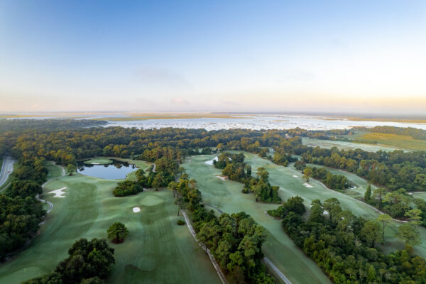 Aerial view of a lush, green golf course surrounded by trees and small ponds, with a road on the left and a distant body of water under a clear blue sky at sunrise.