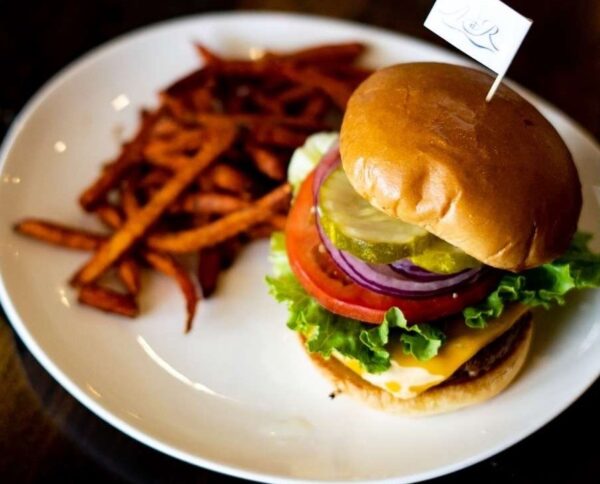 A plate with a cheeseburger topped with lettuce, tomato, red onion, pickles, and a small flag, served next to a side of sweet potato fries.