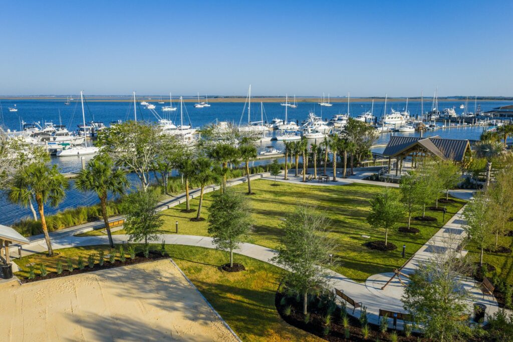 A marina with many boats docked by a park with green lawns, palm trees, walking paths, and a pavilion, all set under a clear blue sky.