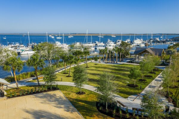A marina with many boats docked by a park with green lawns, palm trees, walking paths, and a pavilion, all set under a clear blue sky.