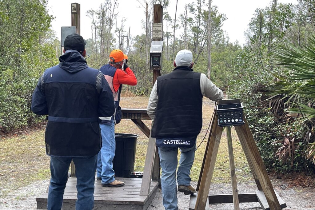 Three people, wearing jackets and jeans, stand at an outdoor shooting range. One person wears an orange cap and adjusts controls, while the others watch. Trees and shrubs surround the area.