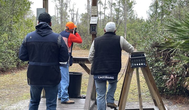 Three people, wearing jackets and jeans, stand at an outdoor shooting range. One person wears an orange cap and adjusts controls, while the others watch. Trees and shrubs surround the area.