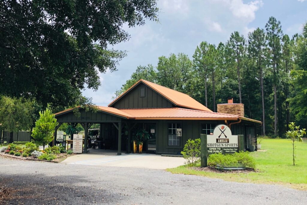 A green building with a brown metal roof stands surrounded by trees and landscaping. A sign in front reads Shotgun Sports Center. The sky is partly cloudy above the rural setting.