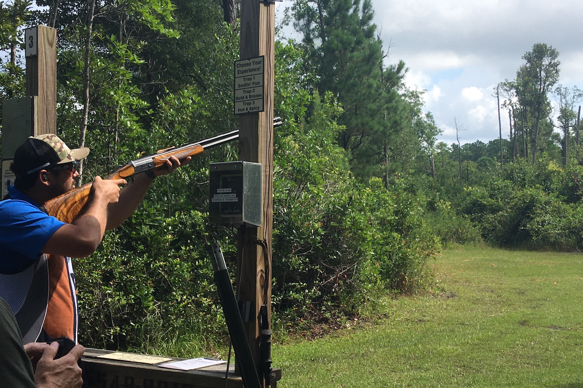 A man wearing sunglasses and a cap aims a shotgun at an outdoor shooting range surrounded by trees and greenery under a partly cloudy sky.