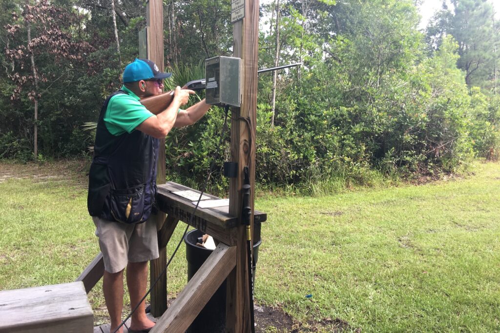 A person wearing a cap and shooting vest aims a shotgun at an outdoor shooting range, standing inside a wooden structure surrounded by green trees and grass.