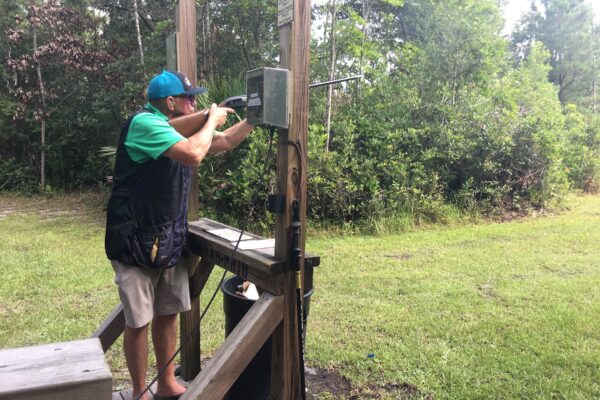 A person wearing a cap and shooting vest aims a shotgun at an outdoor shooting range, standing inside a wooden structure surrounded by green trees and grass.