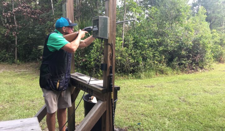 A person wearing a cap and shooting vest aims a shotgun at an outdoor shooting range, standing inside a wooden structure surrounded by green trees and grass.