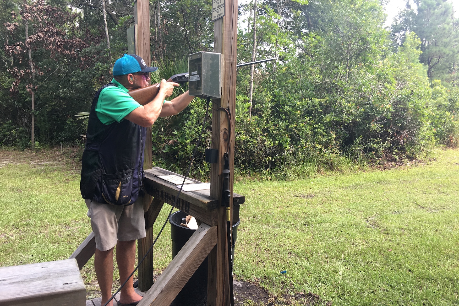 A person wearing a cap and shooting vest aims a shotgun at an outdoor shooting range, standing inside a wooden structure surrounded by green trees and grass.