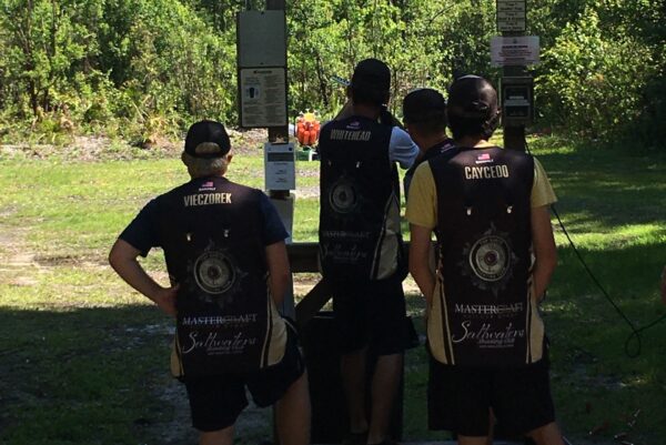 Four people wearing matching shooting team vests stand outdoors at a shooting range, facing away from the camera and looking toward a target area surrounded by trees in sunlight.