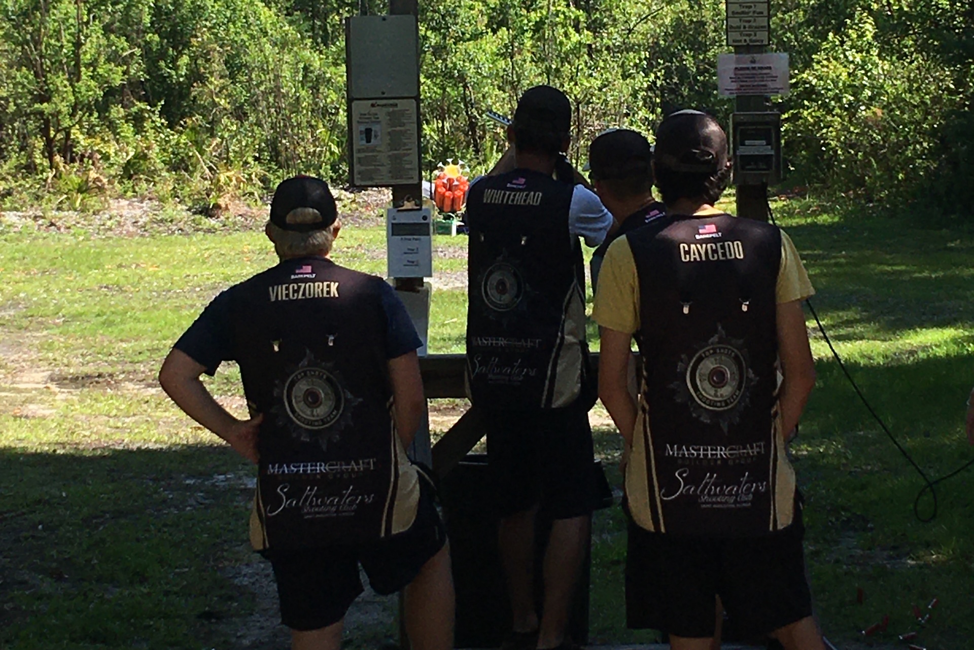 Four people wearing matching shooting team vests stand outdoors at a shooting range, facing away from the camera and looking toward a target area surrounded by trees in sunlight.