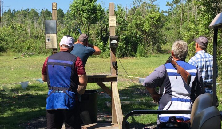 Four people participate in an outdoor skeet shooting activity. One person is aiming a shotgun at clay targets, while the others watch. They stand on grass near wooden platforms, surrounded by trees under a clear sky.