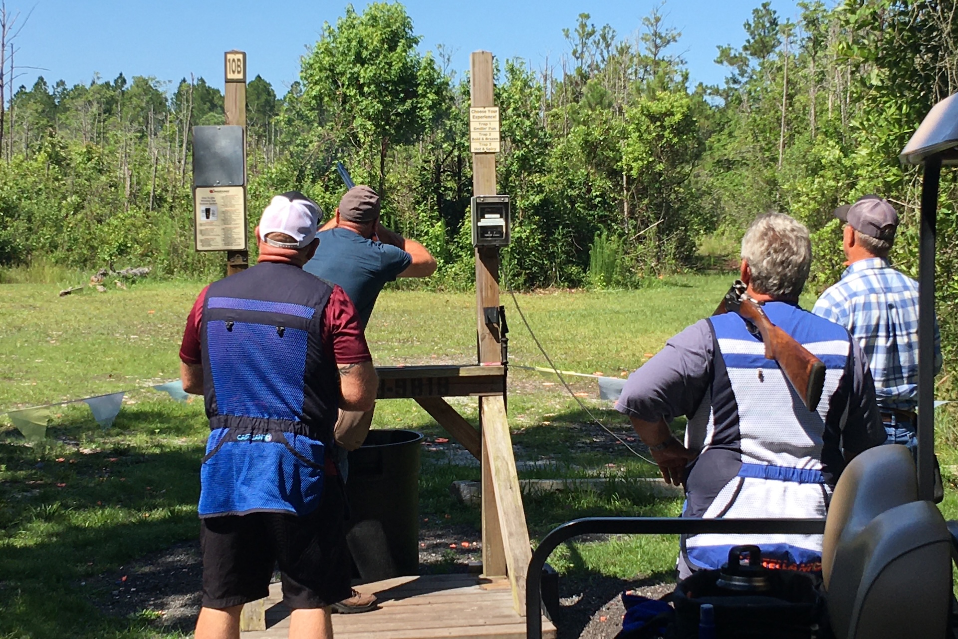 Four people participate in an outdoor skeet shooting activity. One person is aiming a shotgun at clay targets, while the others watch. They stand on grass near wooden platforms, surrounded by trees under a clear sky.