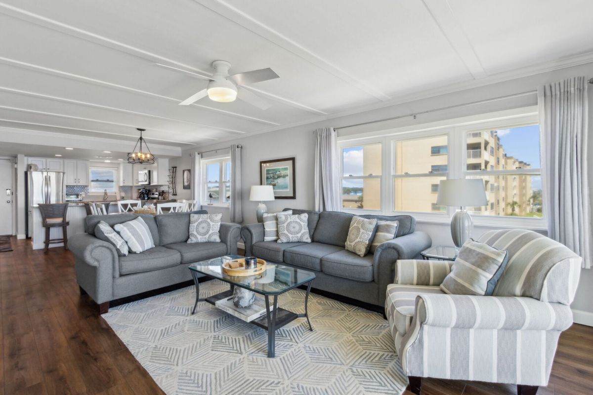 Bright living room with gray sofas, a striped armchair, glass coffee table, and rug, adjoining a kitchen and dining area, large windows offering views of a building and the ocean.