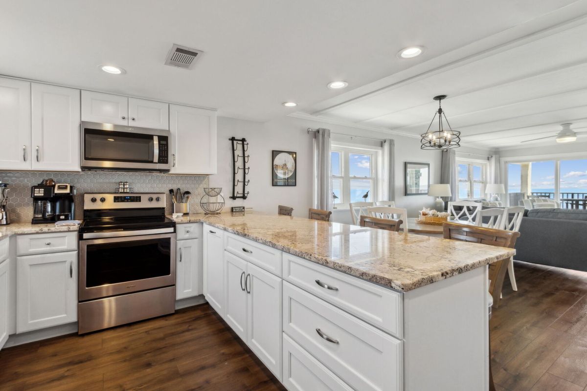 Modern kitchen with white cabinets, stainless steel appliances, and a marble island with barstools. The space opens to a dining and living area with large windows and a view of the water.