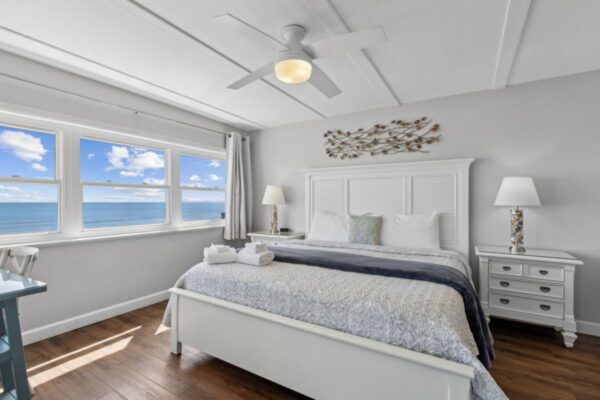 Bright bedroom with a white bed, two bedside tables with lamps, and wall art above the headboard. Large windows offer a scenic ocean view under a blue sky with clouds.