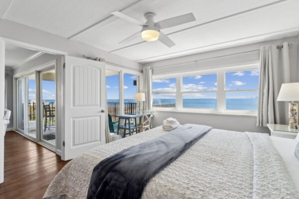 Bright bedroom with large windows, ocean view, a neatly made bed with neutral bedding, bedside lamps, ceiling fan, and door leading to a balcony with patio furniture and clear blue sky above.