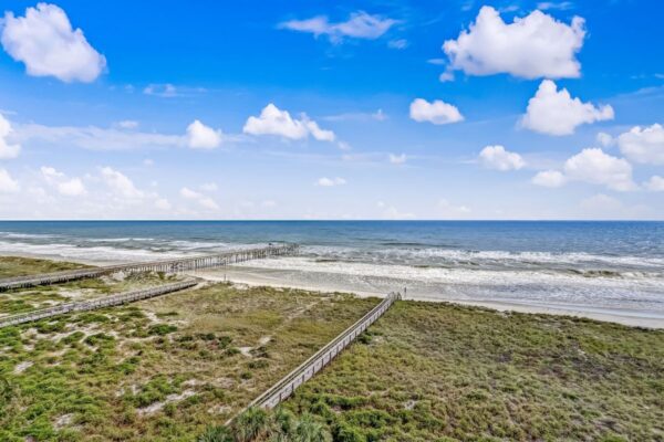 Wooden boardwalks stretch across grassy dunes toward a sandy beach and blue ocean under a bright sky with scattered white clouds.