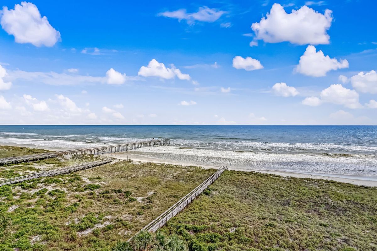 Wooden boardwalks stretch across grassy dunes toward a sandy beach and blue ocean under a bright sky with scattered white clouds.