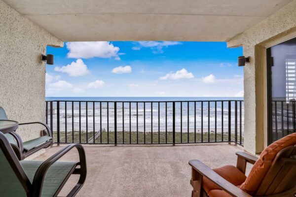 A beachfront balcony with chairs overlooks the ocean, sandy shore, and blue sky with scattered clouds, framed by a black railing and textured walls.