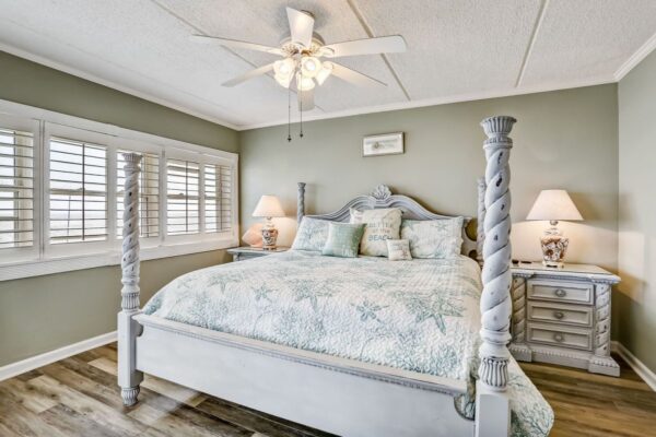 Bedroom with a white four-poster bed, gray-green walls, two matching nightstands with lamps, a ceiling fan, and large shuttered windows. The bedding has a coastal theme in soft pastel colors.