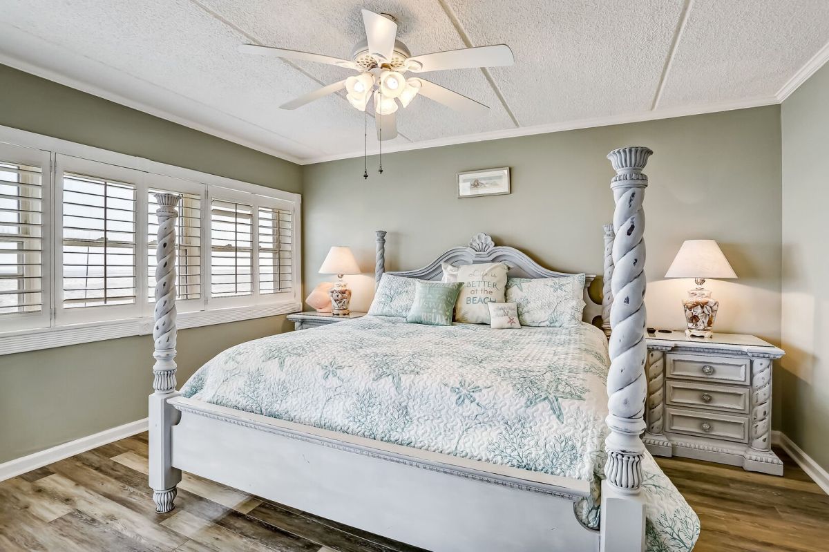 Bedroom with a white four-poster bed, gray-green walls, two matching nightstands with lamps, a ceiling fan, and large shuttered windows. The bedding has a coastal theme in soft pastel colors.