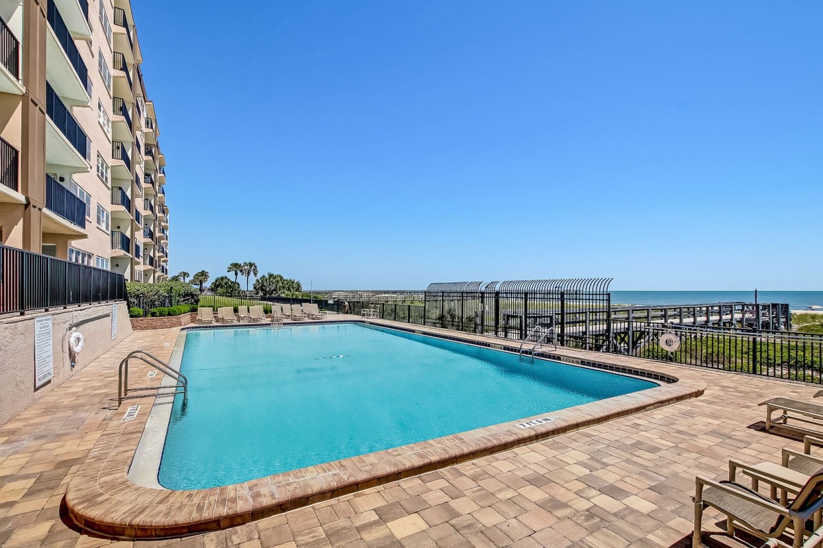 A large outdoor pool with lounge chairs sits beside a multi-story building. The pool overlooks the ocean, palm trees, and a wooden boardwalk under a clear blue sky.