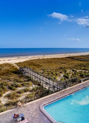 A swimming pool overlooks a sandy beach and ocean under a clear blue sky. A wooden boardwalk stretches over grassy dunes toward the shoreline. Lounge chairs surround the pool area.