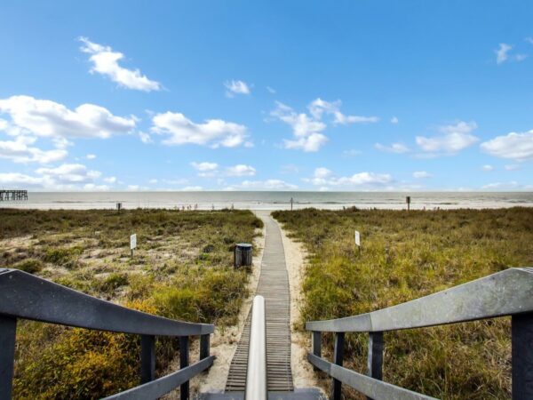 A wooden walkway leads through grassy dunes toward a sandy beach and the ocean, under a mostly clear blue sky with scattered clouds.