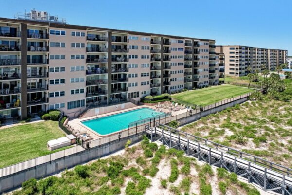 A large apartment complex with balconies overlooks a rectangular outdoor swimming pool, surrounded by a fence, sun loungers, and grassy areas, near a sandy beach walkway under a clear blue sky.