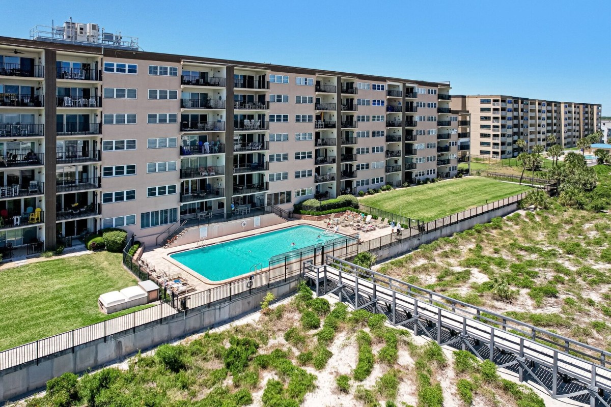 A large apartment complex with balconies overlooks a rectangular outdoor swimming pool, surrounded by a fence, sun loungers, and grassy areas, near a sandy beach walkway under a clear blue sky.