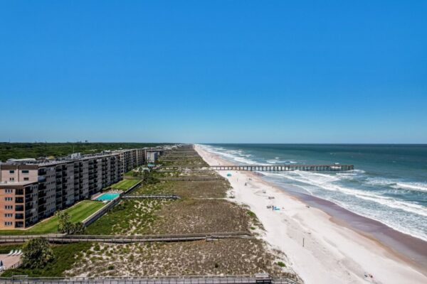 Aerial view of a sandy beach with ocean waves, seaside buildings, a long pier extending into the water, and a clear blue sky. Few people can be seen on the beach.