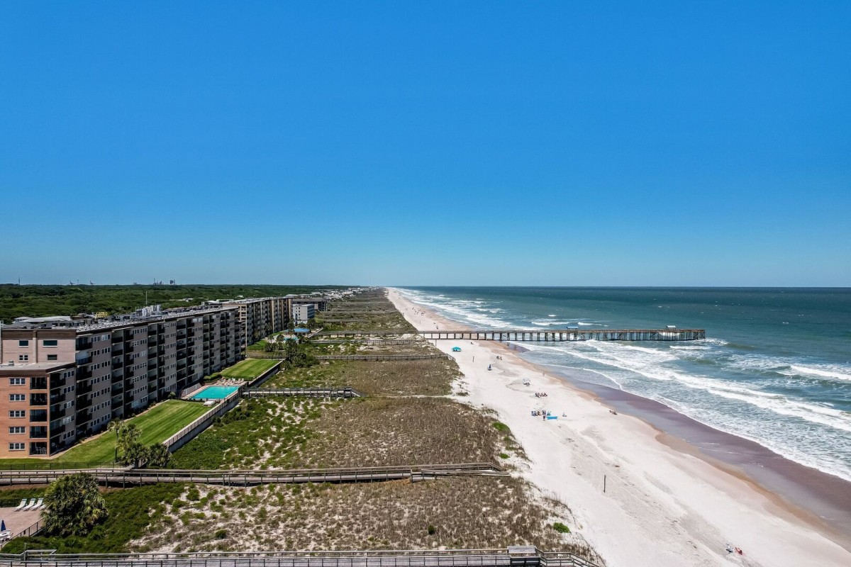 Aerial view of a sandy beach with ocean waves, seaside buildings, a long pier extending into the water, and a clear blue sky. Few people can be seen on the beach.