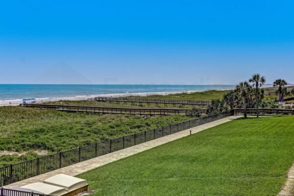 View of a grassy lawn bordered by a black fence, leading to a sandy beach with boardwalks and palm trees under a clear blue sky. The ocean is calm, and a few umbrellas are visible on the shore.